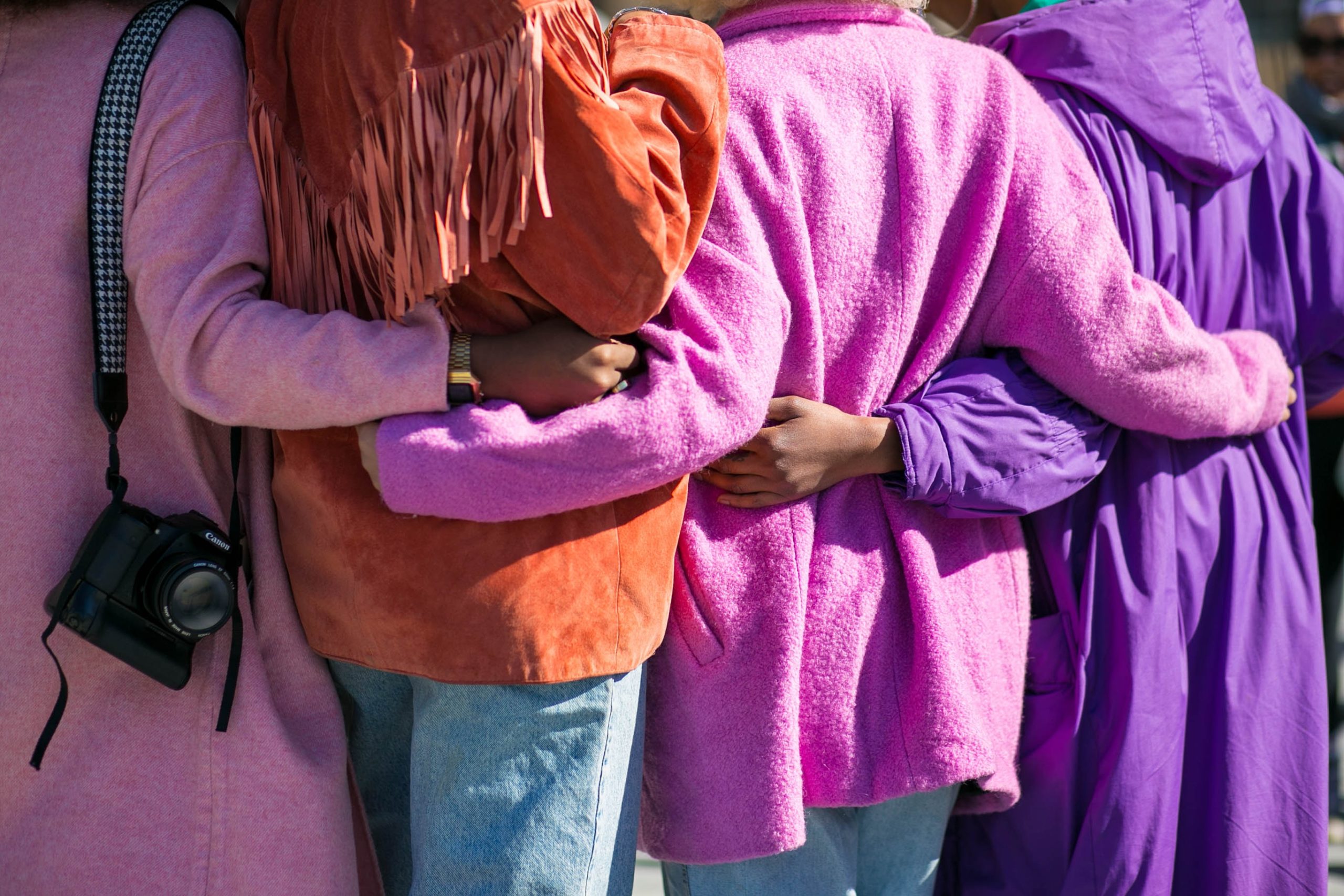 4 individuals facing away from the camera wearing multicolored coats with their arms interlocked at the waist