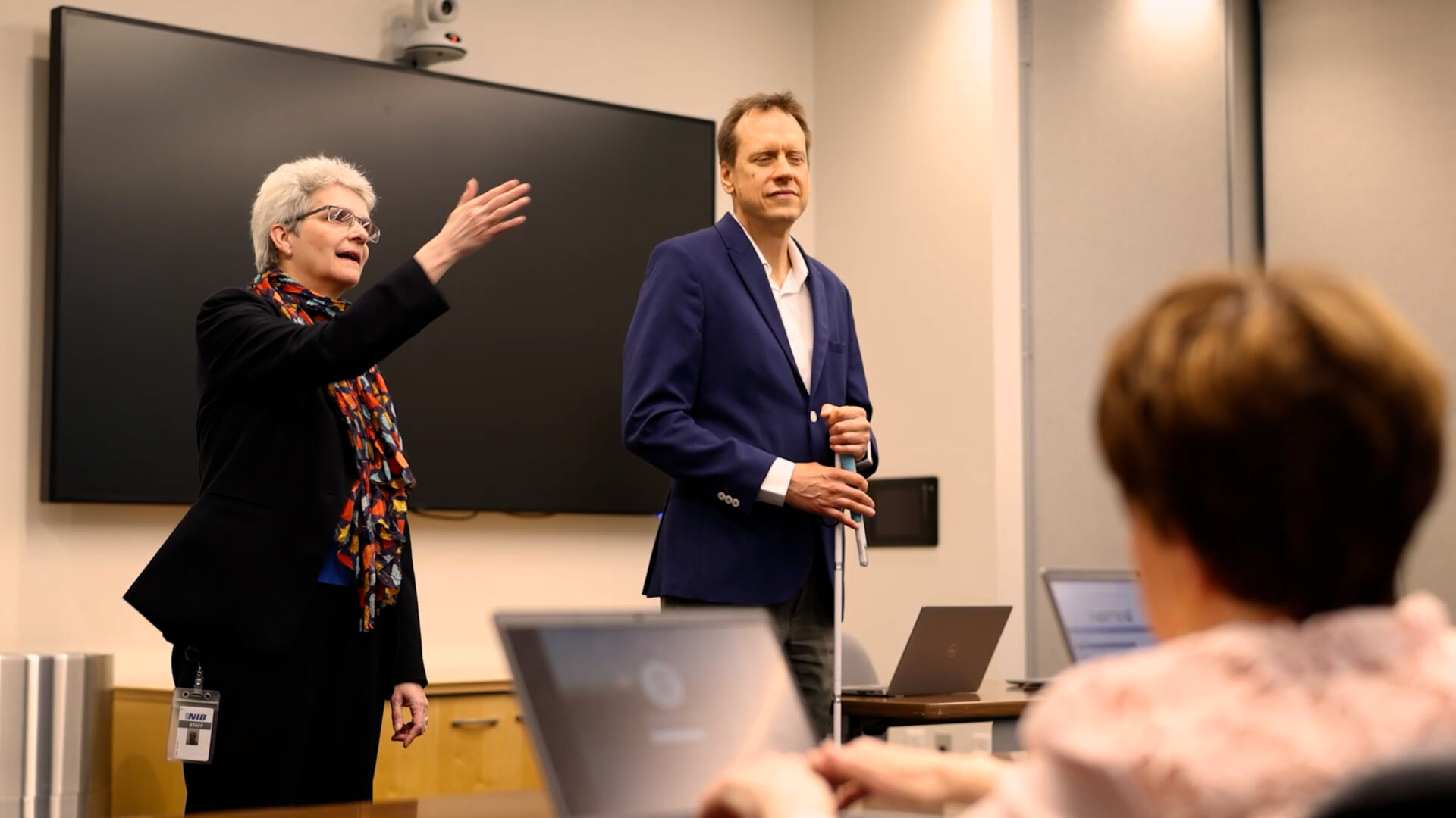 A man and woman stand in front of a classroom of adult students. The woman is speaking to the group.