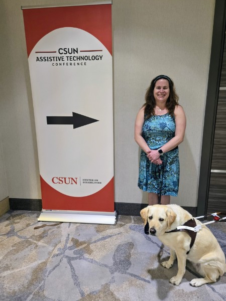 A woman and her guide dog stand near a conference sign.